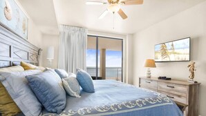 Ein Schlafzimmer mit einem großen Bett, einem Deckenventilator, Blick aufs Meer durch einen Balkon und einem Fernseher auf einer Holzkommode.