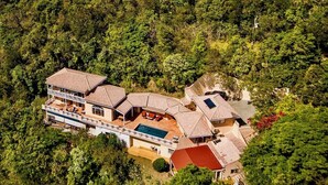 Une grande maison à plusieurs étages, dotée d’une piscine et entourée d’une forêt dense.
