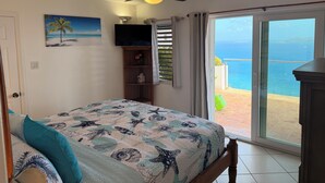 Une chambre à coucher avec un lit, un ventilateur de plafond, une télévision, un tableau, une étagère et une vue sur l’océan.