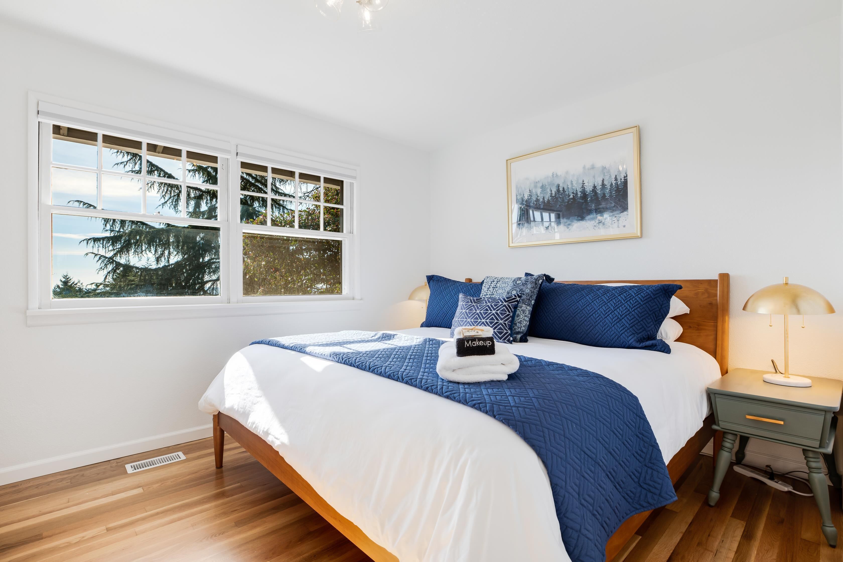 A bedroom with a large bed, a nightstand, a window with a view of trees, and a framed picture on the wall.