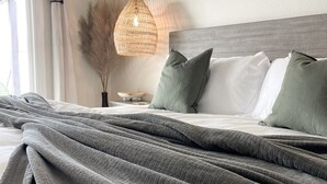 Un lit bien fait, recouvert d’une couverture grise, une suspension lumineuse en fibres tressées et un vase contenant des plantes séchées sur une table de chevet.