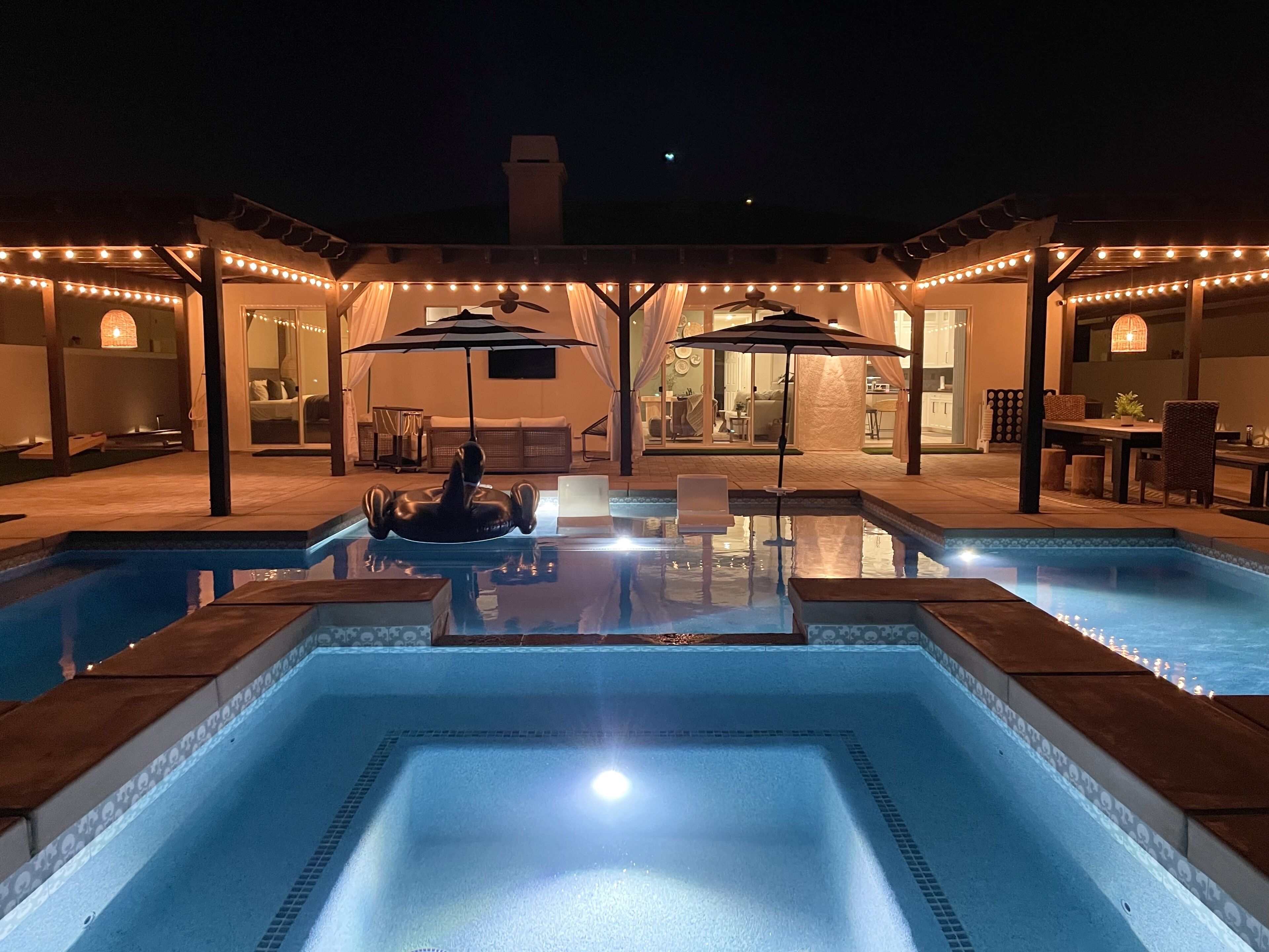 A swimming pool with two lounge chairs and umbrellas, surrounded by a covered patio area with string lights.