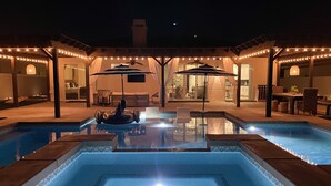 Une piscine avec deux chaises longues et des parasols, entourée d’une terrasse couverte agrémentée de guirlandes lumineuses.