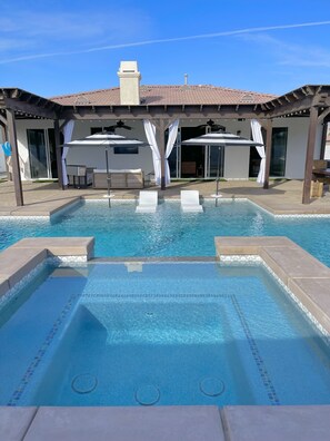 A rectangular swimming pool with clear blue water, surrounded by a pergola with white curtains and umbrellas.