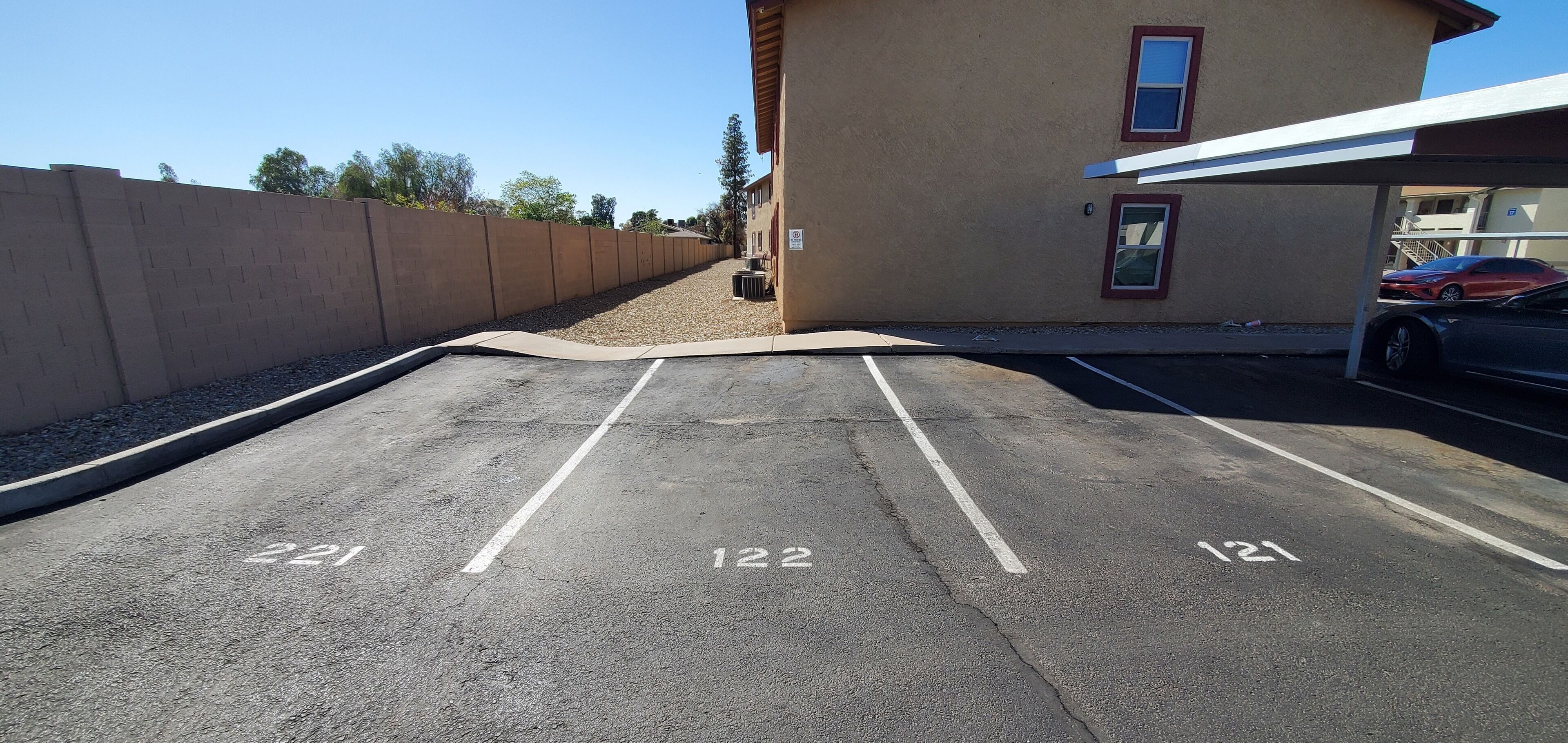 A parking lot with three marked spaces, a building with a red door, and a car parked under a covered area.