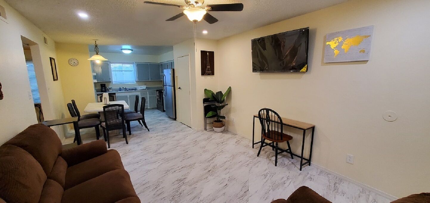 A living room with a ceiling fan, a dining area with chairs and a table, and a kitchen in the background.