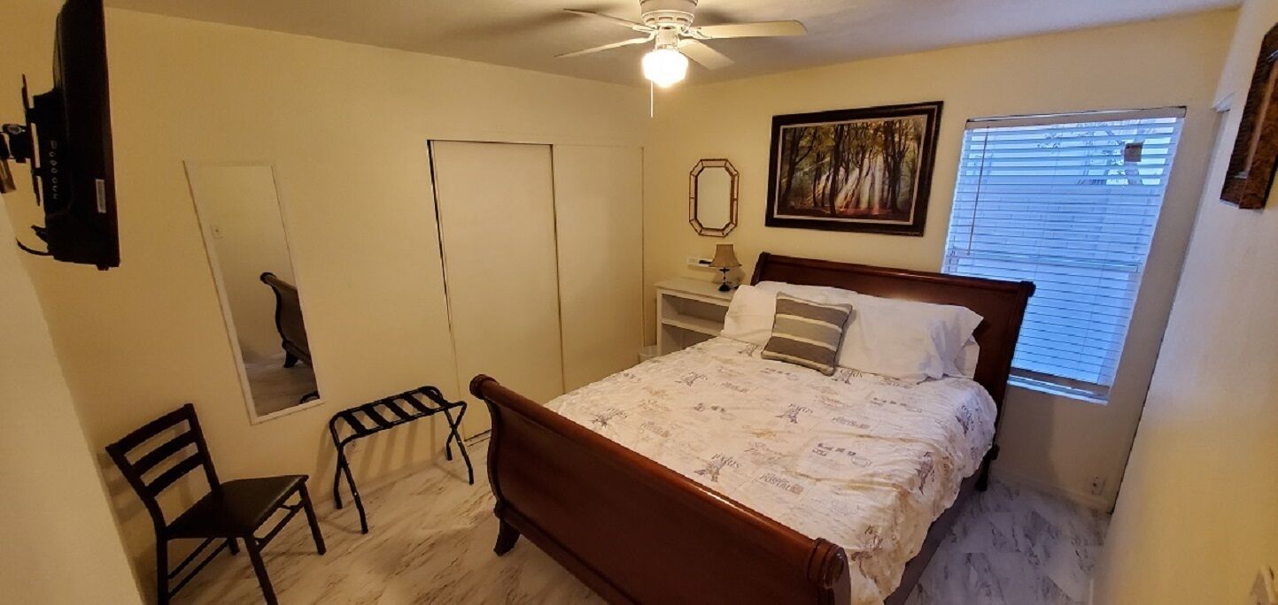 A bedroom with a bed, a chair, a desk, a ceiling fan, a window with blinds, and a door.