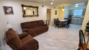 Un salon avec un canapé marron, un coin repas avec une table et des chaises, et une cuisine à l’arrière-plan.
