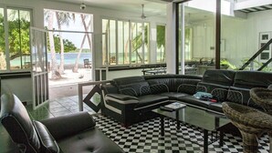 A modern living room with a black leather sofa, a glass coffee table, and wicker chairs. Large windows offer a view of the beach and palm trees.