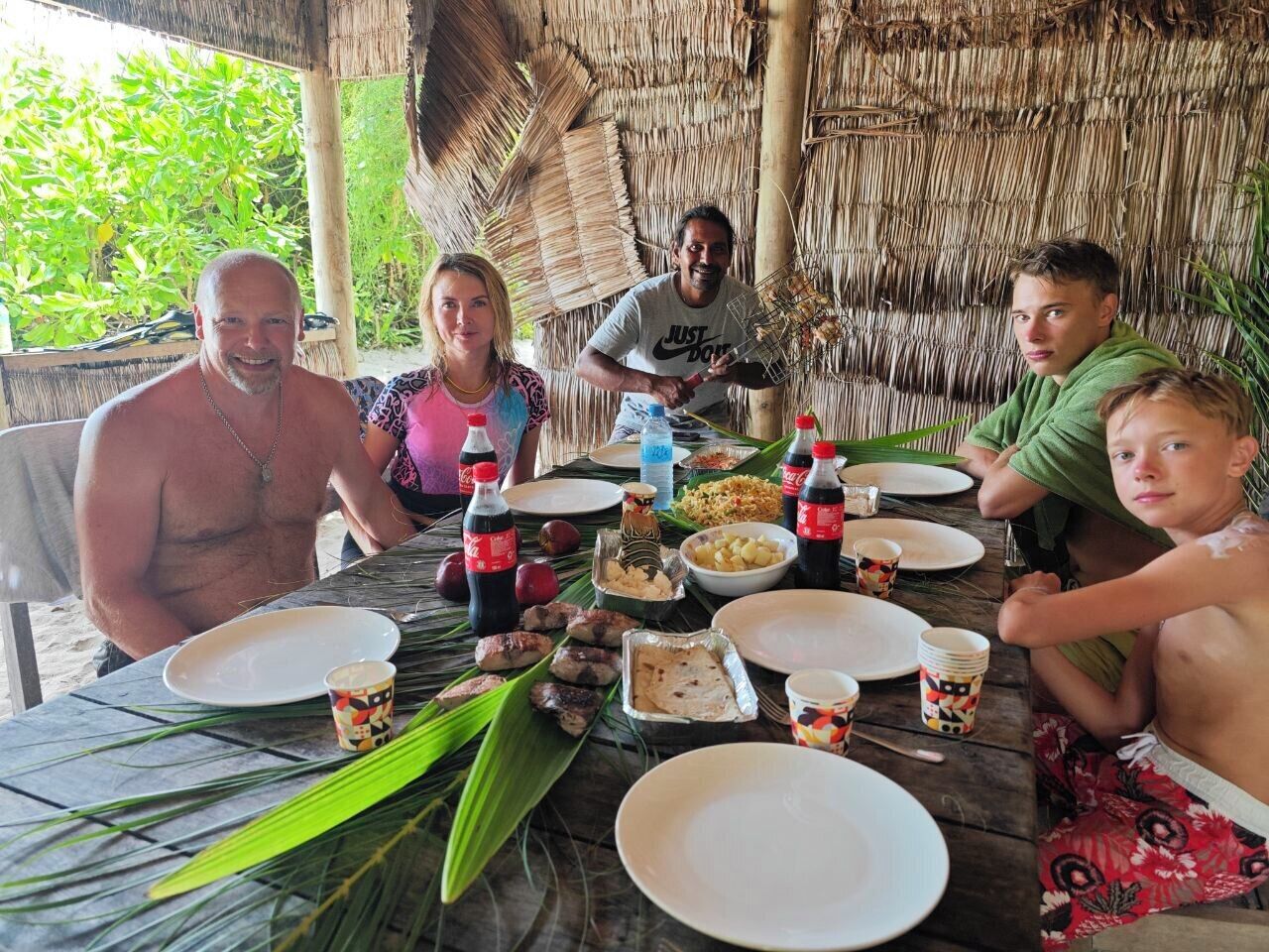 A group of people dining at an outdoor table with a thatched roof, surrounded by tropical plants.