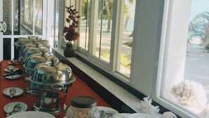 A buffet setup with a variety of food items and a juice dispenser, set against a backdrop of large windows overlooking a beach.