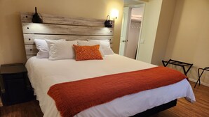 A neatly made bed with white bedding and a red throw blanket, a wooden headboard, a bedside table with a lamp, and a closet with hanging clothes.