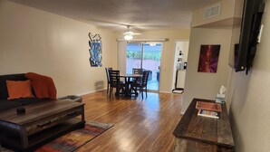 A living room with a dark wooden coffee table, a sofa with orange pillows, a dining area with a table and chairs, and a kitchen in the background.