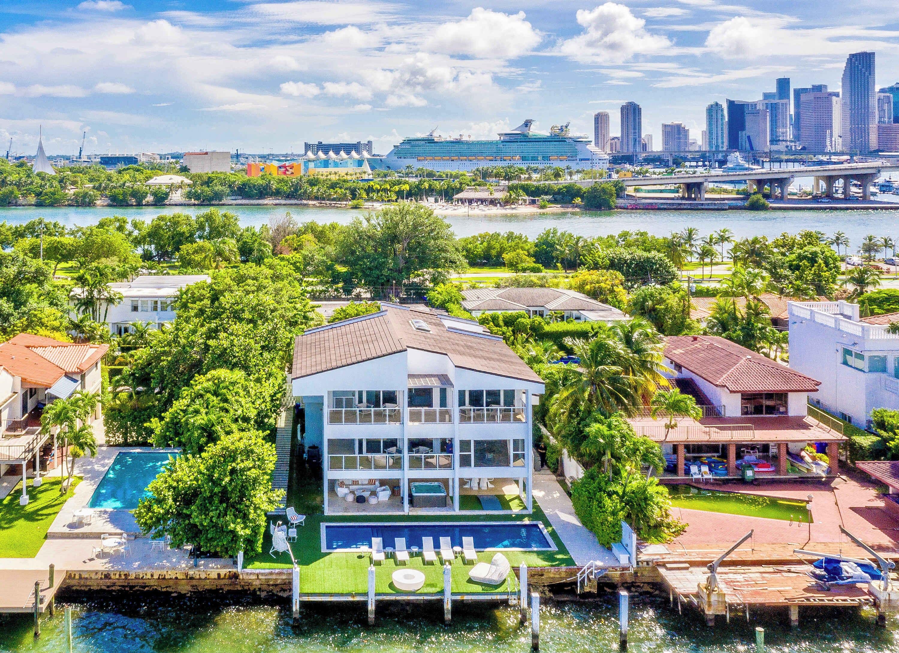 Aerial view of a three-story waterfront villa featuring a private swimming pool, outdoor lounge areas, private dock, landscaped grounds, multiple balconies, and direct water access, with bay and city skyline views visible from the property.