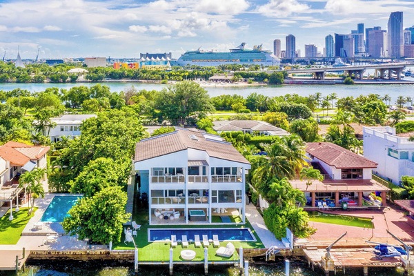 Aerial view of a three-story waterfront villa featuring a private swimming pool, outdoor lounge areas, private dock, landscaped grounds, multiple balconies, and direct water access, with bay and city skyline views visible from the property.