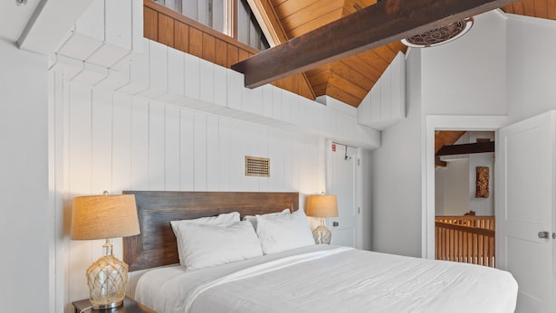 A bedroom with a large bed, two bedside tables with lamps, a wooden headboard, and a white ceiling.