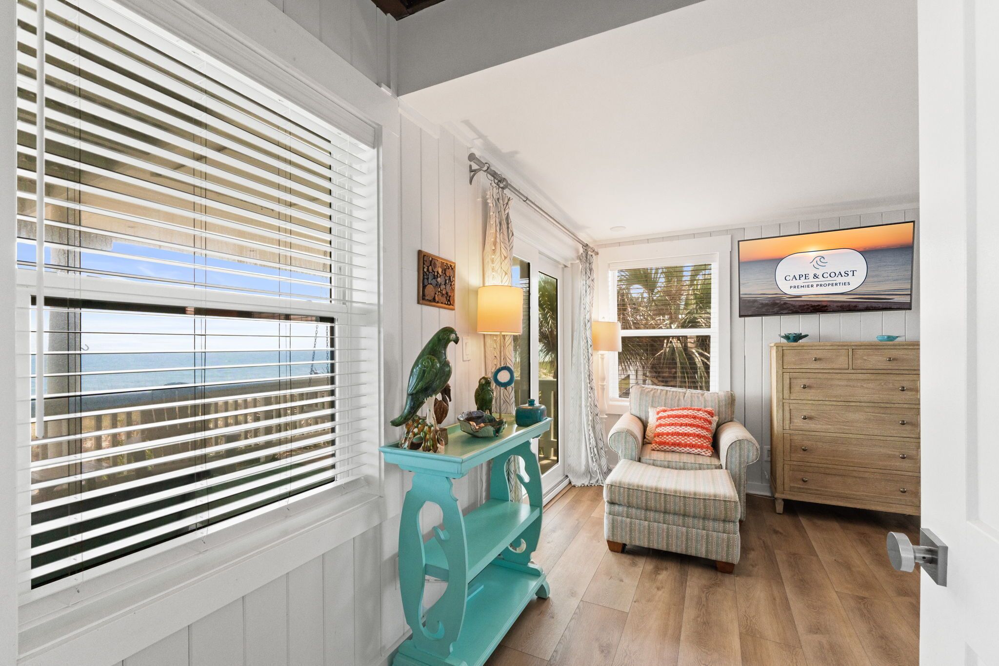 Una habitación con vistas al mar a través de amplios ventanales, un aparador turquesa con objetos decorativos, un sillón con reposabrazos y un plaid estampado, y un armario de madera.