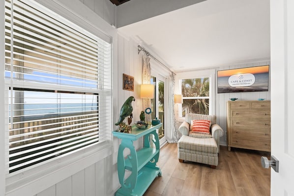 Quarto com vista para o mar através de grandes janelas, uma mesa de consola verde-água com objetos decorativos, uma poltrona estofada com mantas estampadas e um armário de madeira.