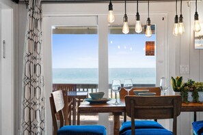 A dining area with a view of the ocean, a table set with plates and glasses, and hanging light fixtures. - South Wind: Gorgeous Beachfront Home, Private Pool, Hot Tub, Fire Pit (St. George Island)