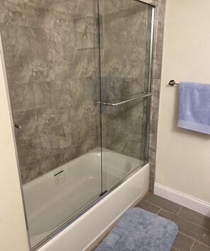 A glass-enclosed shower with a bathtub, grey stone walls, and a blue towel on a rack.