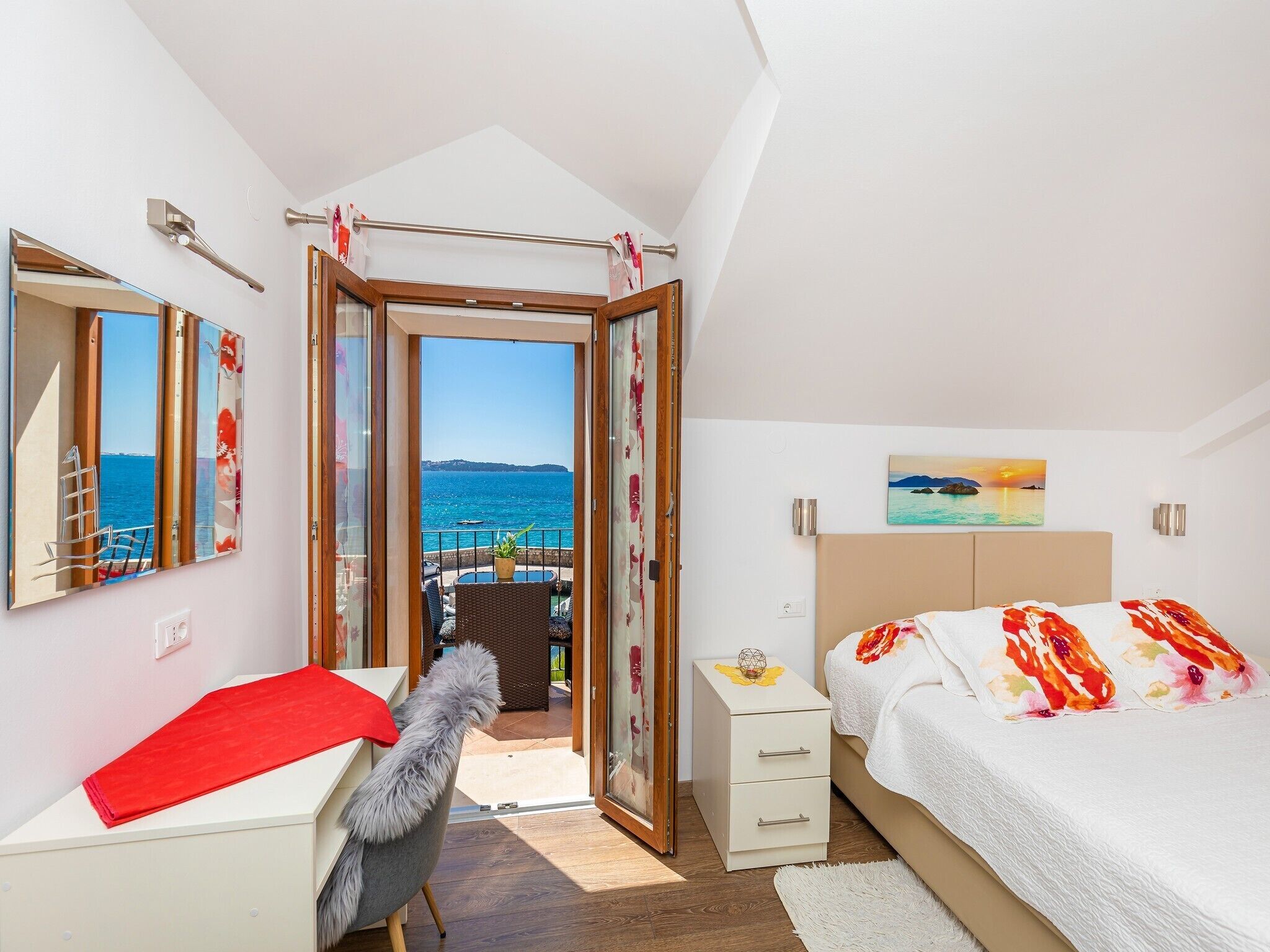 A bedroom with a bed, a desk, a chair, a nightstand, and a view of the sea.