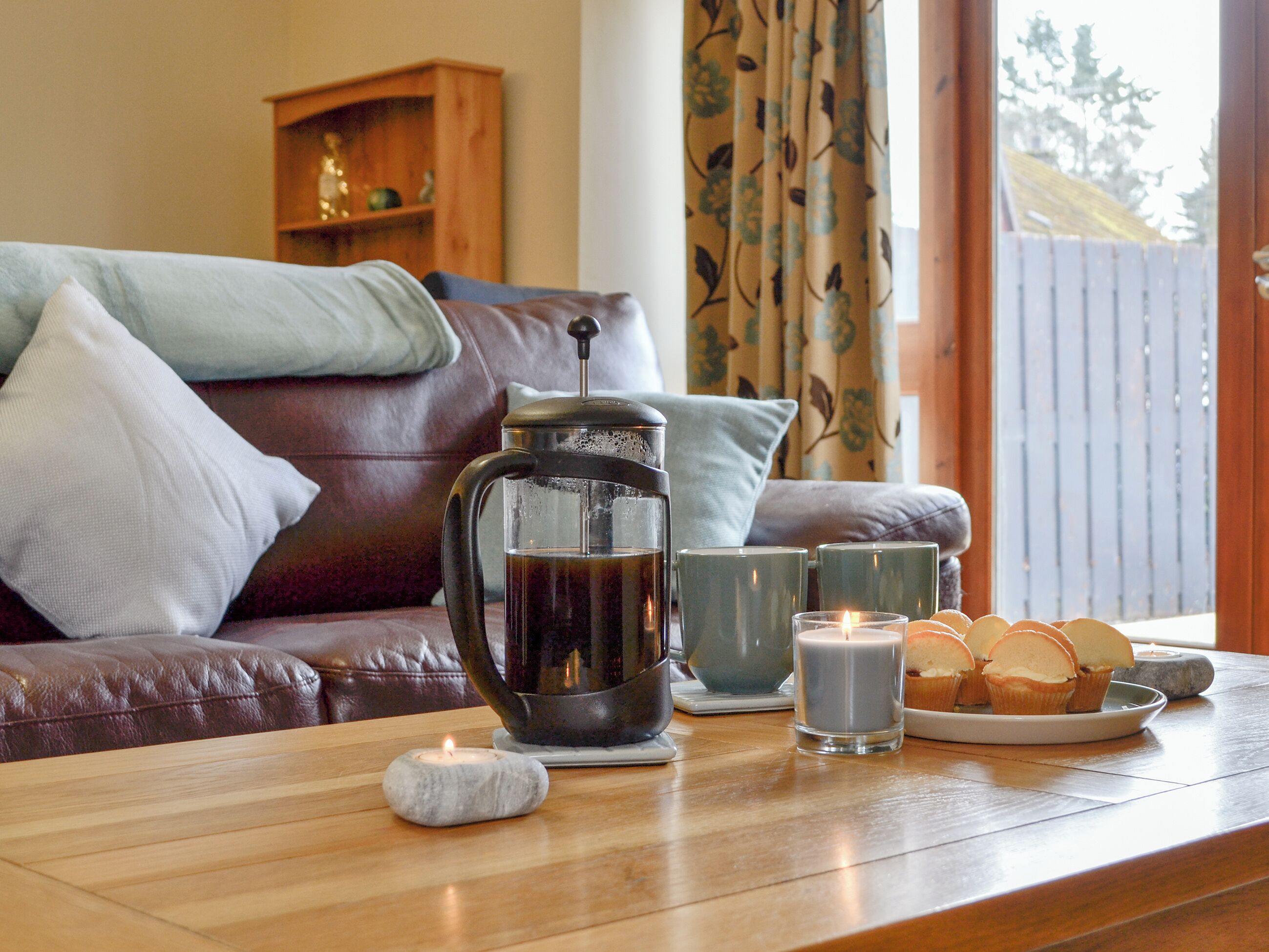 A cozy living room with a coffee pot, candles, and a plate of muffins on a wooden table.
