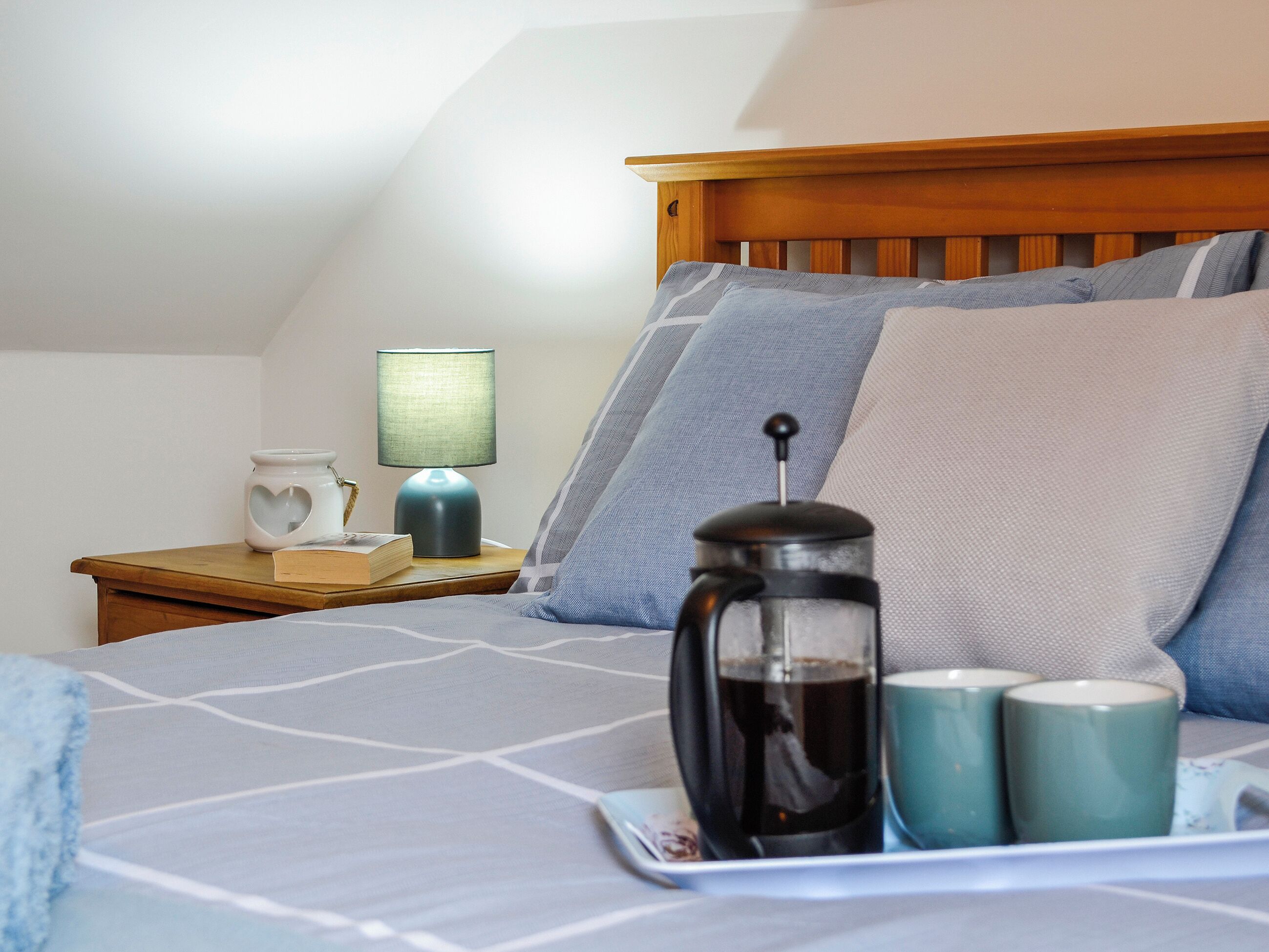 Un lit bien fait, recouvert d’une couvre-lit à motifs géométriques, une table de chevet avec une lampe, une machine à café et deux tasses.