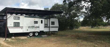 Una caravana blanca aparcada bajo una estructura con techo, rodeada de árboles y un campo cubierto de hierba.