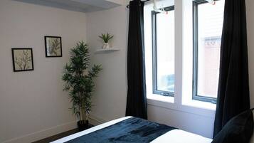 Un dormitorio con una cama, una ventana con cortinas negras, una planta en maceta y dos cuadros en la pared.