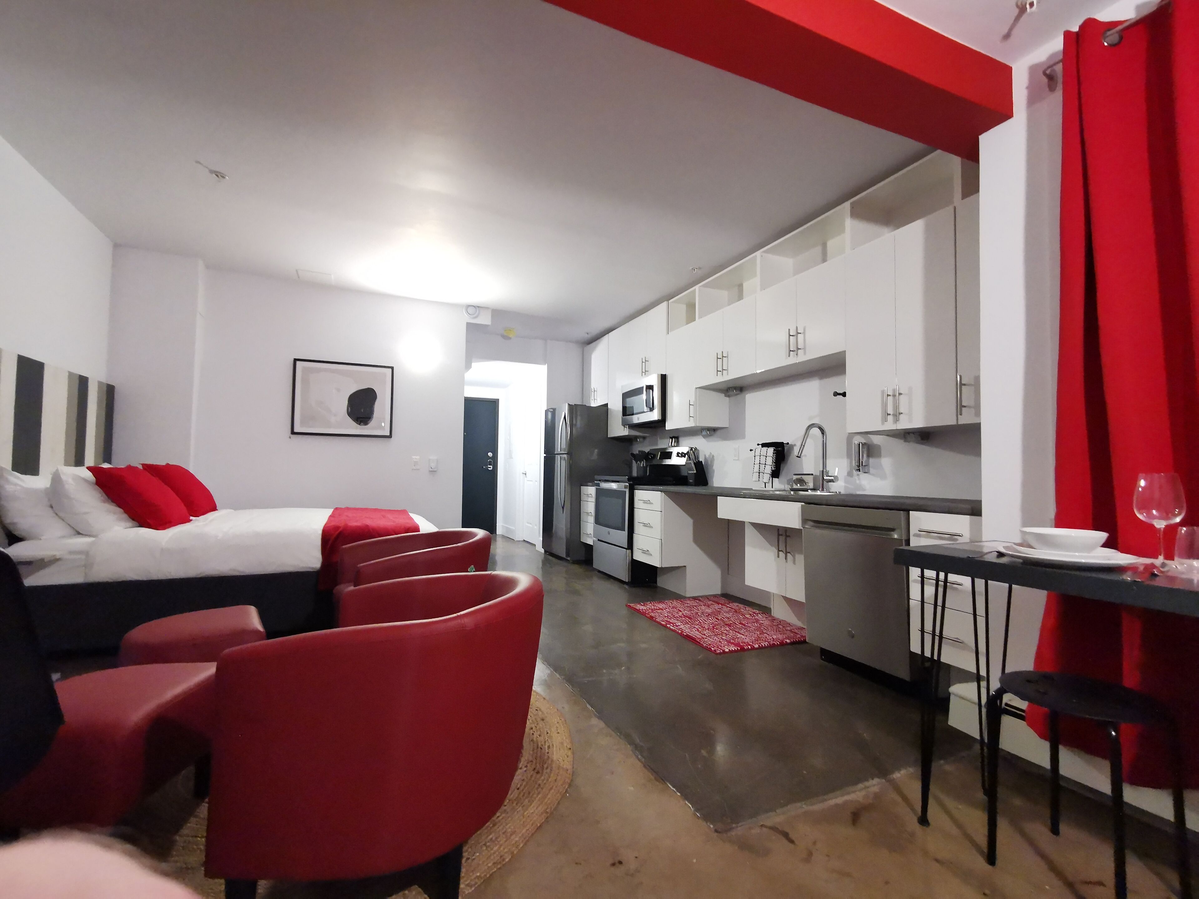 A modern hotel room with a bed, red chairs, a small kitchen, and a dining area.
