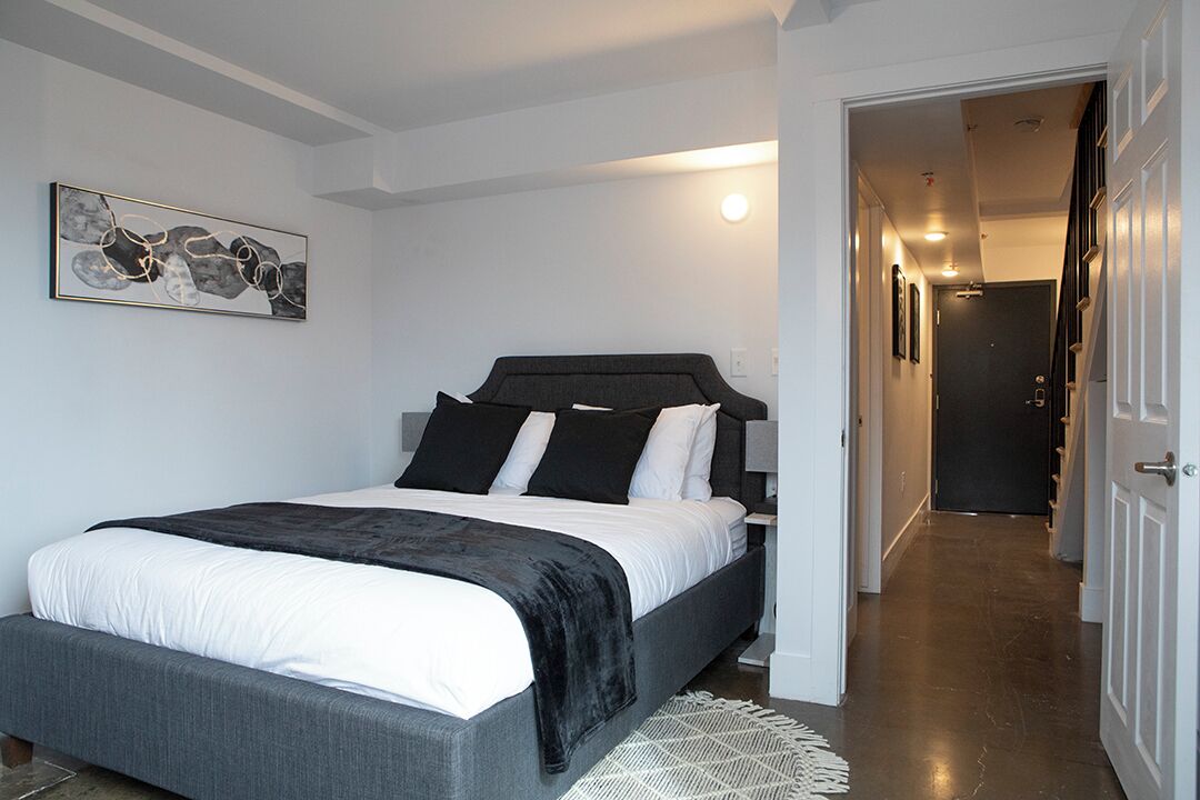 A modern bedroom with a large bed, a dark headboard, and a white wall with a framed abstract painting.