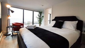 Una habitación de hotel moderna con una cama grande, una silla roja, una mesita pequeña y un amplio ventanal con vistas a la ciudad.