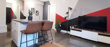 Un salon moderne avec une table basse en bois, deux fauteuils marron, un téléviseur et un coin cuisine doté d’un comptoir-bar.