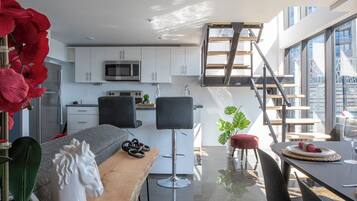 Een moderne keuken met witte kastjes, een magnetron en een fornuis. Er is een eethoek met een tafel, stoelen en een rode poef. Een trap leidt naar een hoger gelegen verdieping.