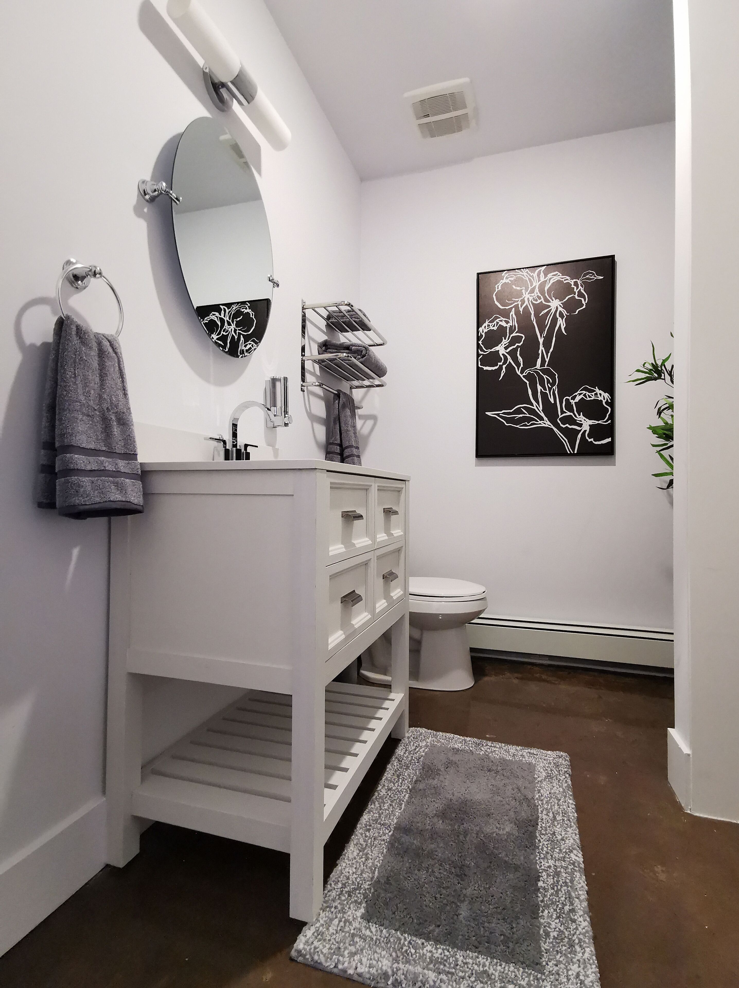 Une salle de bain moderne avec un meuble-lavabo blanc, un miroir rond, des toilettes et une œuvre d’art florale en noir et blanc accrochée au mur.