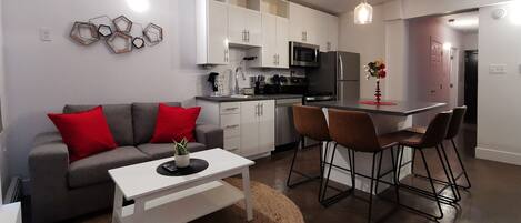 Un espacio habitable compacto con un sofá gris, cojines rojos, una mesa de centro blanca y una zona de cocina con armarios blancos, microondas y refrigerador.