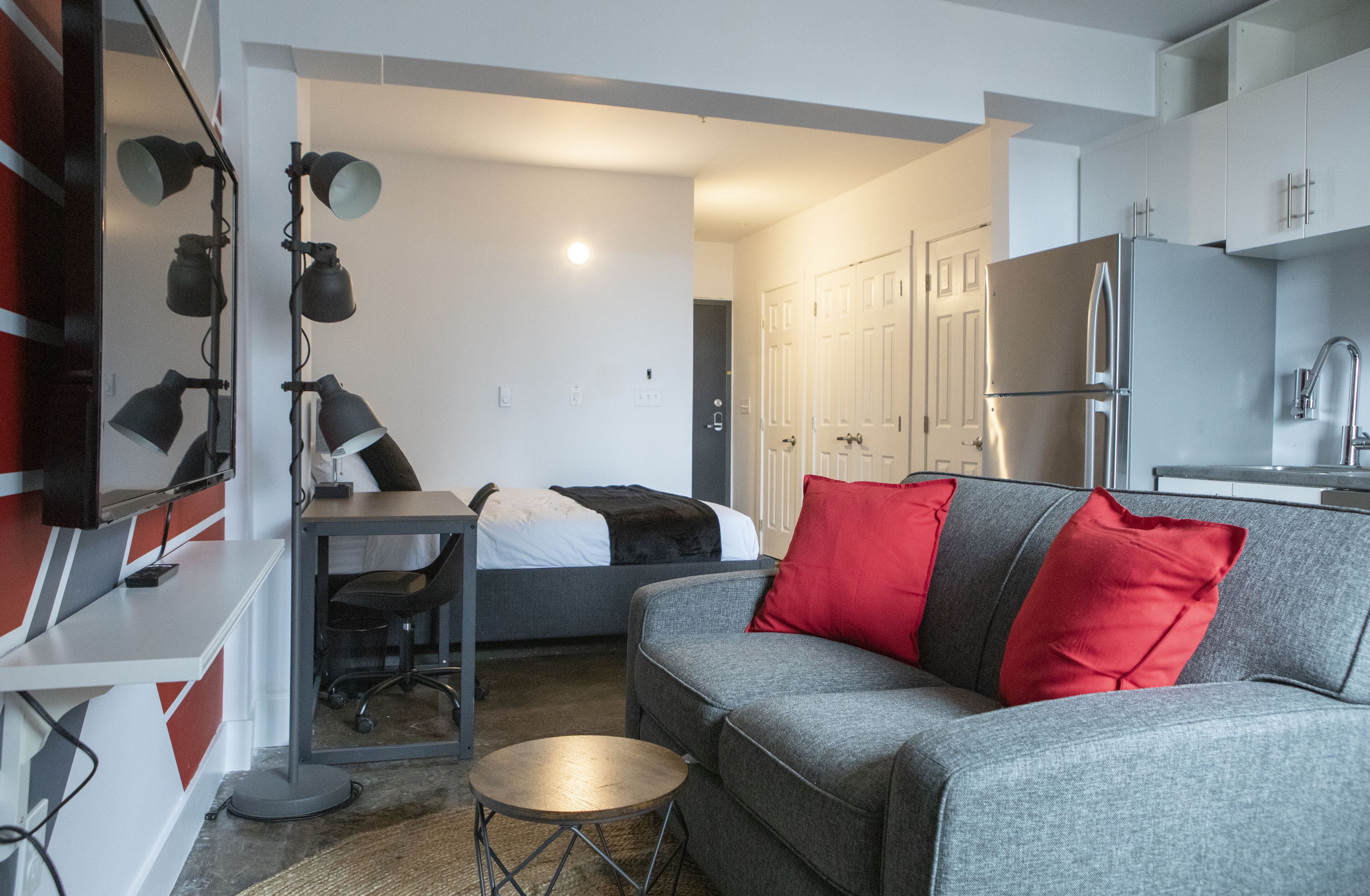 Een compacte woonruimte met een grijze bank, rode en zwarte kussens, een klein rond tafeltje, een bureau met stoel, en een kitchenette met een koelkast.