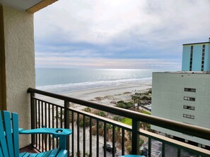 Un balcón con vista a la playa y al océano, un estacionamiento y un edificio con múltiples ventanas.