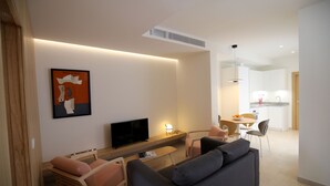 Een moderne woonkamer met een flatscreen televisie, een bank, een salontafel en een eethoek, met een keuken op de achtergrond.