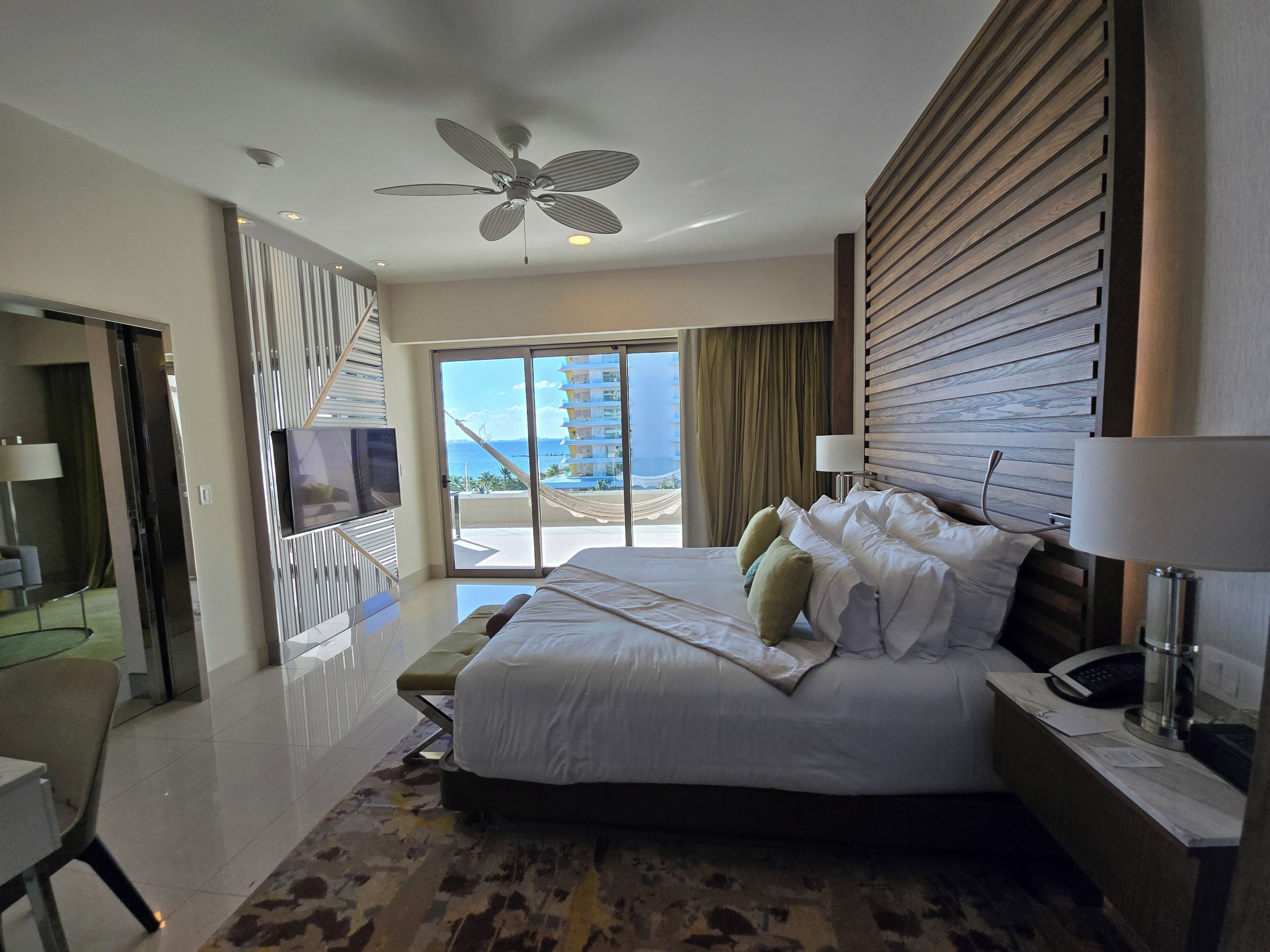 A modern hotel room with a large bed, a television, a desk with a lamp, and a view of the ocean.