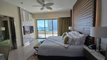 A modern hotel room with a large bed, a television, a desk with a lamp, and a view of the ocean.