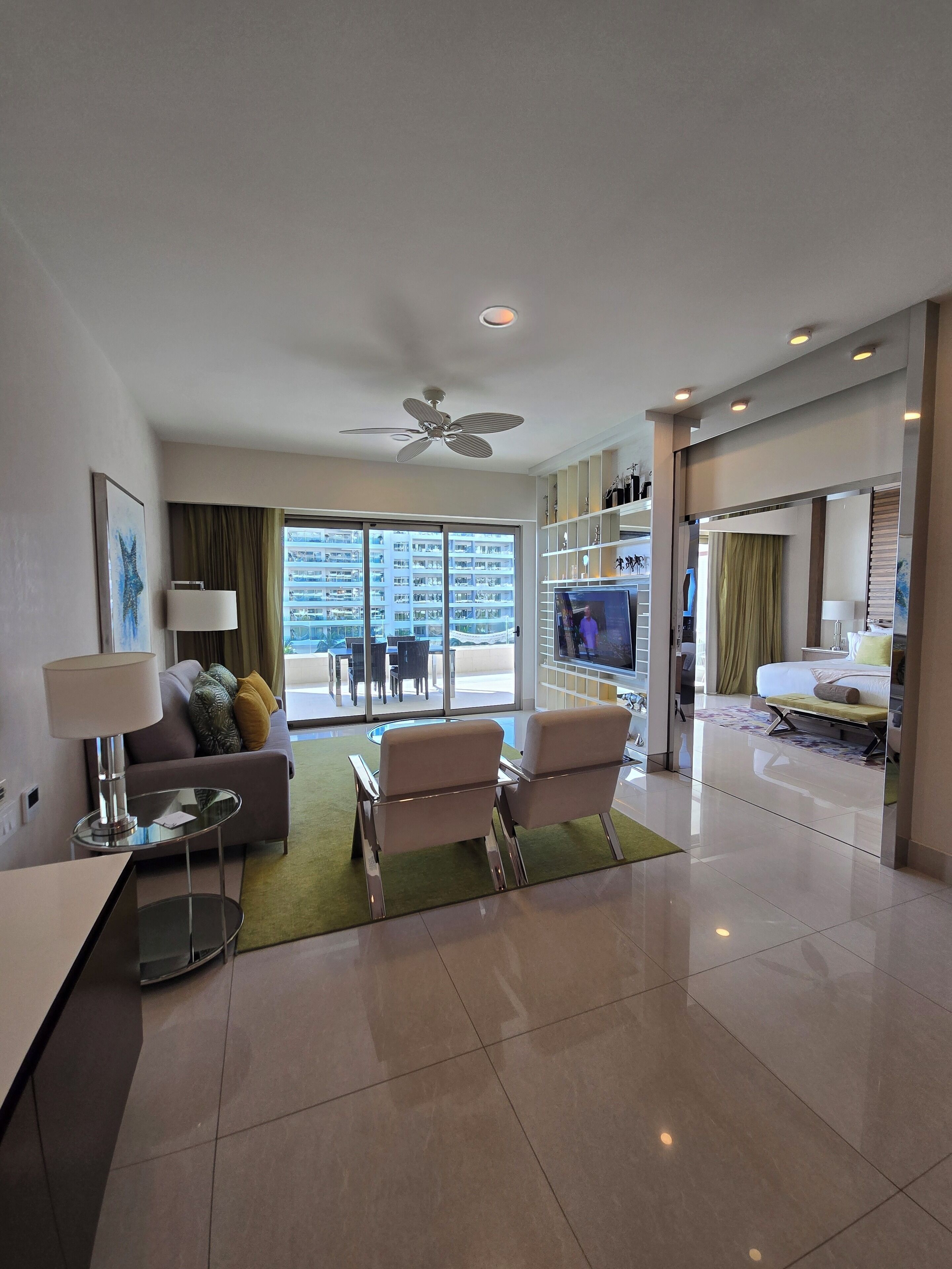 A modern hotel room with a balcony, a sofa, two armchairs, a TV, and a dining area with a table and chairs.
