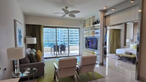 Ein modernes Hotelzimmer mit Balkon, einer Sitzecke, zwei Sesseln, einem Fernseher und einem Essbereich mit Tisch und Stühlen.