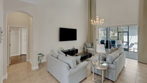 Ein großzügiges Wohnzimmer mit einer großen Couch, einem Couchtisch und einem an der Wand montierten Fernseher. Eine Chandelier ist vorhanden und es gibt einen Blick ins Freie durch große Fenster.