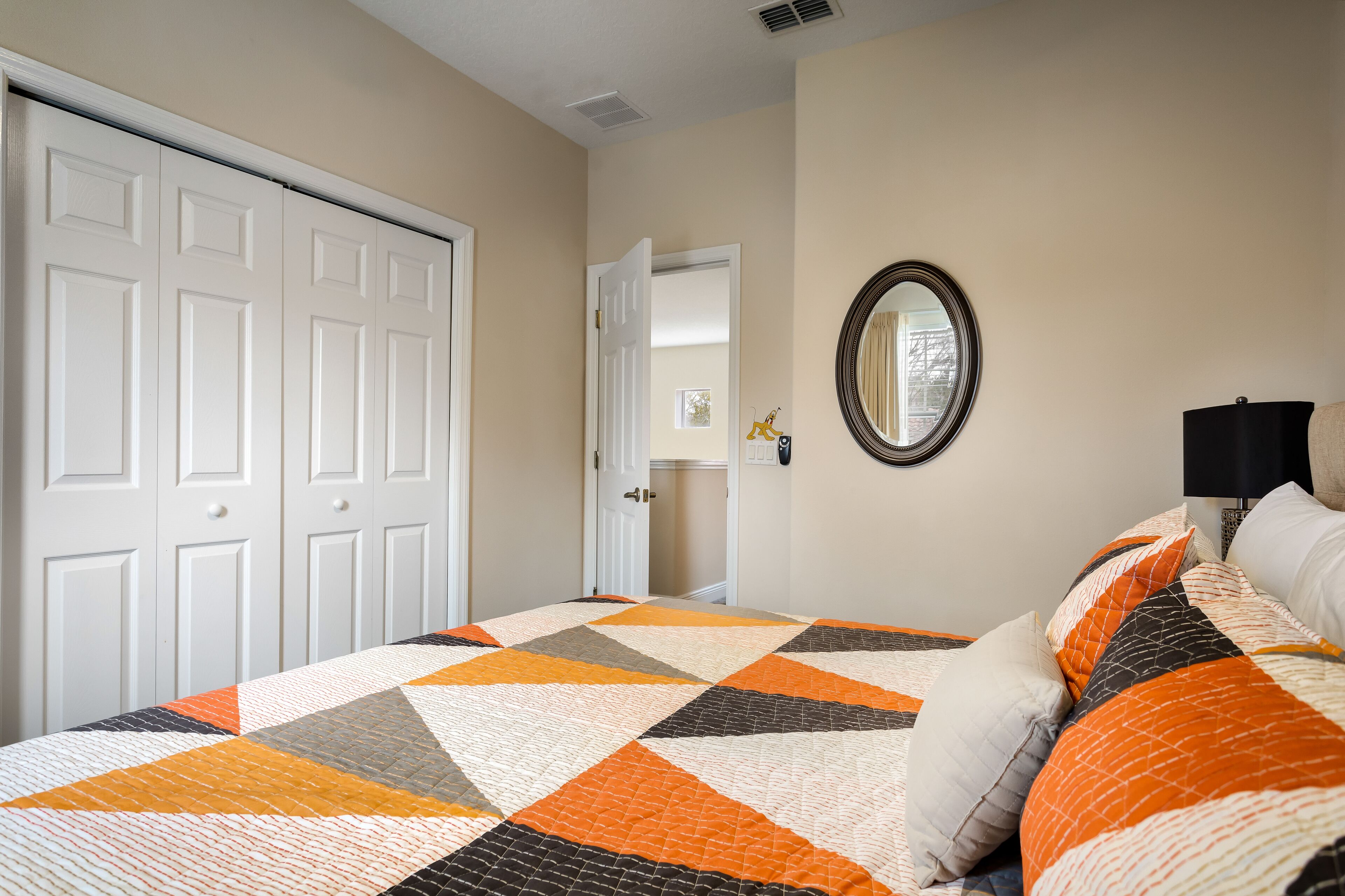 A bedroom with a bed, a door, a mirror, and a nightstand with a lamp.