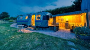 A stationary RV with a kitchen area and outdoor seating, adjacent to a wooden building with a warm interior light. - Airstream, Devon Hideaways -  a shepherds hut that sleeps 2 guests  in 1 bedroom (Kenn)