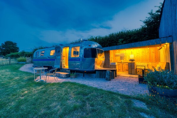 A stationary RV with a kitchen area and outdoor seating, adjacent to a wooden building with a warm interior light. - Airstream, Devon Hideaways - a shepherds hut that sleeps 2 guests in 1 bedroom (Kenn)