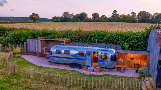 A vintage camper parked in a rural setting with a wooden cabin and a gravel driveway. - Airstream, Devon Hideaways -  a shepherds hut that sleeps 2 guests  in 1 bedroom (Kenn)