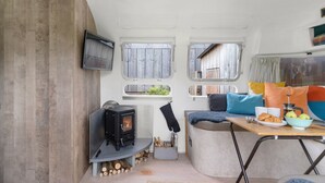 A cozy interior with a wood-burning stove, a dining table with a bowl of fruit, and a comfortable seating area. - Airstream, Devon Hideaways -  a shepherds hut that sleeps 2 guests  in 1 bedroom (Kenn)