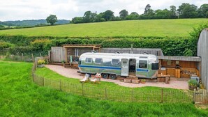 A camper parked in a rural setting with a wooden structure and a fenced garden area. - Airstream, Devon Hideaways -  a shepherds hut that sleeps 2 guests  in 1 bedroom (Kenn)